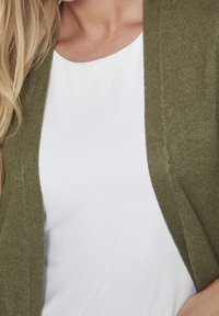 Olivgrön cardigan med öppen front och mjuk textur, överlagd på en vit topp. Slät stickad material och minimal detaljering.