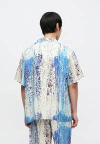 Camisa de manga corta en tela ligera con un patrón abstracto en azul y crema. Presenta salpicaduras notables de azul y púrpura con un ajuste holgado.