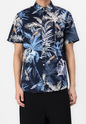 Mand iført navyblå kortærmet skjorte med knapper og tryk af tropiske palmeblade og kolibri, kombineret med sorte bukser.