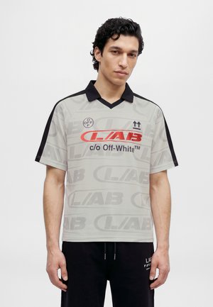 Un mannequin masculin se tient devant un fond uni, vêtu d’un polo gris clair à motif graphique, avec un col noir et un pantalon noir.