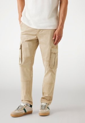 Beige cargobroek met zijzakken, taps toelopende pasvorm, katoenen materiaal, gestikte details, gedragen met grijze en groene sneakers.