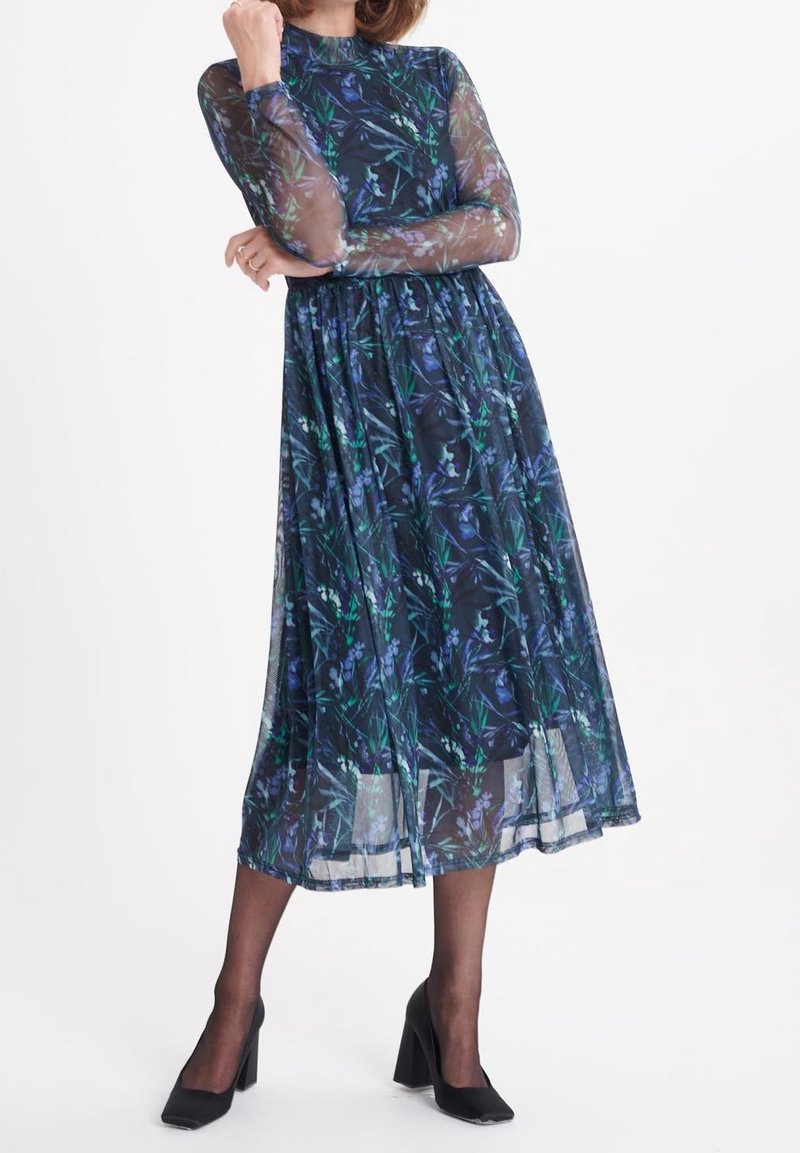 Femme portant une robe midi à manches longues, bleu marine à motifs floraux avec des manches transparentes et des chaussures noires à talons blocs, debout.