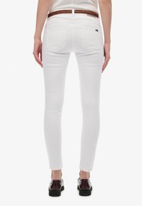 Jeans blancos ajustados con una textura suave, que cuentan con dos bolsillos traseros y un cinturón trenzado marrón. Llevados con zapatos negros.