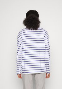 Persona con capelli ricci che indossa una camicia bianca a maniche lunghe con strisce orizzontali blu e pantaloni grigio chiaro, vista da dietro.