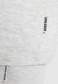 Lichtgrijze stof met een close-up van een shirtnaad en een zwart label met de tekst "SMILODOX". De textuur lijkt zacht en iets gemêleerd.