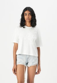 Witte katoenen t-shirt met korte mouwen, met textuur en bloemenborduursel aan de linkerkant. Gepaard met versleten denim shorts.