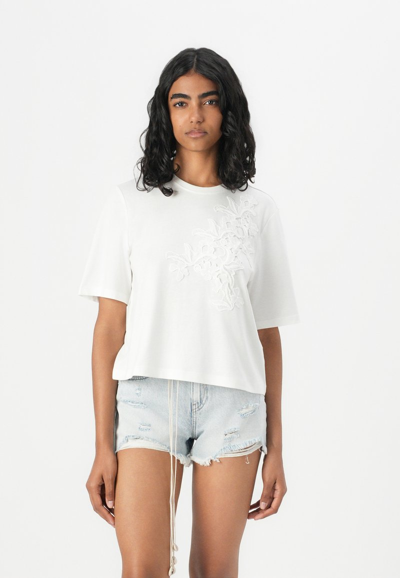 Witte katoenen t-shirt met korte mouwen, met textuur en bloemenborduursel aan de linkerkant. Gepaard met versleten denim shorts.
