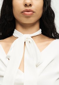 Γυναίκα με μεσαίο τόνο δέρματος που φοράει λευκό μπλουζάκι με δεμένο φουλάρι στο λαιμό, δείχνοντας τα χείλη, το πηγούνι, τον λαιμό και τις κλείδες.