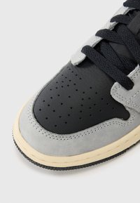 Grå mockaskinn och svart läder sneakers med perforeringar, platta svarta snören och en gummisula med en ljusbeige kant.