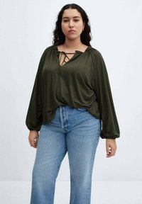 Bluză lungă cu mânecă, de culoare verde măsliniu, cu decolteu legat cu șnur și umeri plisati, asortată cu blugi evazați de culoare albastru deschis. Material moale cu cădere.