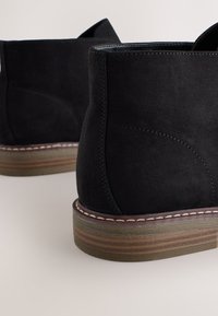 Bottines en daim noir avec bout rond, accents cousus et semelle en caoutchouc superposée marron et verte. Talon plat, design minimaliste.