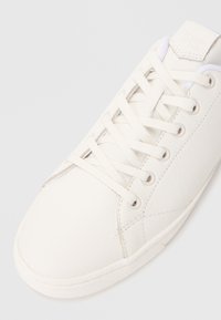 Baskets blanches en cuir avec une surface texturée, bout rond, lacets blancs plats et détails de couture subtils. Design minimaliste, sans logos visibles.