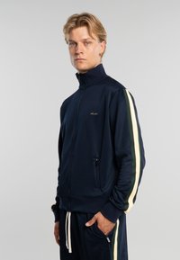 Joven con chándal azul marino con franjas crema y verde oscuro, de pie contra un fondo gris liso, con la mano en el bolsillo, mirando hacia adelante.