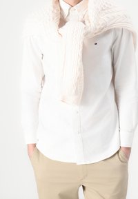 Biała koszula z guzikami w połączeniu z kremowym swetrem dzianym zarzuconym na ramiona. Beżowe spodnie dopełniają stylizację.