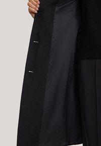 Zwarte coatinterieur met een zachte textuur en satijnen afwerking. Met zilverkleurige hardware, twee binnenzakken en op maat gemaakte randen.