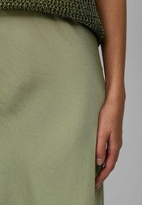 Un tissu texturé vert forme un vêtement fluide, complété par un haut tricoté de couleur vert foncé. Une main repose sur la couture latérale de la jupe.