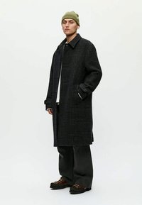 Jeune personne portant un bonnet vert, un long manteau à carreaux foncés, un pantalon large noir et des chaussures marron, se tenant devant un fond clair.