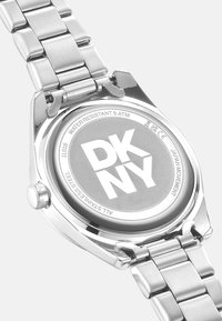 Zilveren roestvrijstalen horloge met een geborstelde afwerking, grijze wijzerplaat met "DKNY" erop en een schakelen bandontwerp; waterbestendig tot 5 ATM.