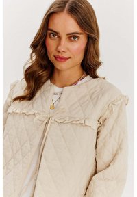 Beżowa pikowana kurtka z falbankami, okrągłym dekoltem i zawiązywaniem. Tkanina ma miękką teksturę i subtelny wzór w romby.