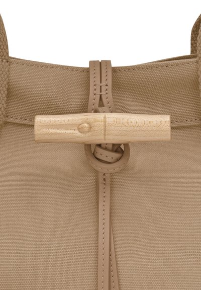 Beige canvas bag with a textured finish, featuring a wooden toggle closure and leather accents, emphasizing simplicity and functional design.
