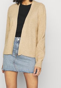 Beige ribbad cardigan med öppen front, långa ärmar och avslappnad passform, i kombination med en ljusblå denimkjol.