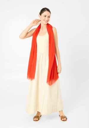 Frau trägt ein ärmelloses cremefarbenes Kleid, einen leuchtend roten Schal über den Schultern drapiert, braune Sandalen, lächelnd und berührt ihren Hals.