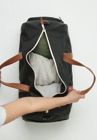 Svart duffelväska med bruna läderhandtag, uppzipad för att visa vikta kläder, inklusive grå och gröna plagg inuti.