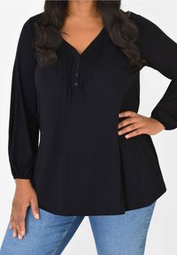 Svart blus med långa ärmar, V-hals, veck vid halsringningen och knäppning med knapp. Mjuk tyg, avslappnad passform, kombinerad med jeans.