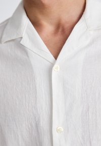 Chemise en lin blanc avec un col en V, une texture subtile et une fermeture à un bouton, mettant en avant son design léger et respirant.