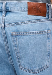 Baklomme og midjebånd på lyseblå denimjeans med brun skinnpatch med Calvin Klein-logo på midjebåndet.