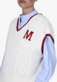 Witte kabelgebreide vest met een rode V-hals en armsgaten, voorzien van een rode "M" borduursel op de voorkant, gedragen over een lichtblauw overhemd met kraag.