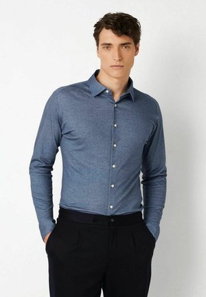 Junger Mann mit dunklem, lockigem Haar, der ein blaues, langärmeliges Hemd mit Knöpfen und schwarze Hose trägt, steht mit den Händen in den Taschen.