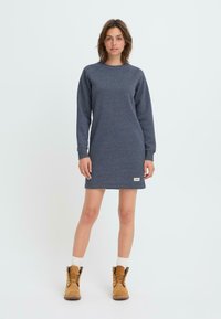 Robe-sweat à manches longues, de couleur gris foncé, avec un col rond et des poignets côtelés, arborant une petite étiquette sur l'ourlet. Associée à des bottes beiges.