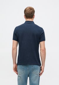 Ναυτικό μπλε πόλο μπλουζάκι με κοντά μανίκια, φτιαγμένο από υφαντό βαμβάκι, με κλασικό γιακά και ίσιο στρίφωμα. Φοριέται με ανοιχτόχρωμο τζιν παντελόνι.