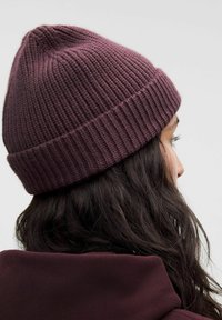 Persona con cabello largo y oscuro, llevando un gorro de punto acanalado de color púrpura oscuro y una chaqueta a juego de color púrpura oscuro, visto de lado y por detrás.