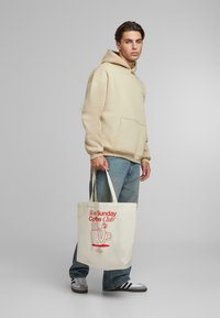 Beige hoodie med en frontlomme, parret med lyseblå jeans. Hvid tote bag med rød grafisk tekst: "Slow Sunday Coffee Club."