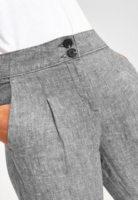 Grijze linnen broek met een getailleerde pasvorm, voorzien van twee zwarte knopen op de taille, voorplooien en zijzakken voor extra detail.