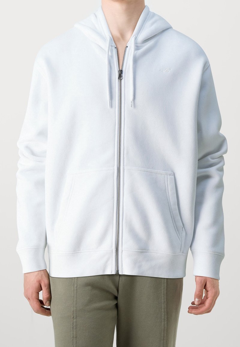 Bílá mikina na zip vyrobená z měkkého materiálu, s kapucí na stahování, předními kapsami a žebrovanými manžetami. Minimalistický design bez vzorů.