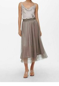 Femme portant un top sans manches en satin à col drapé, une jupe midi plissée avec une superposition transparente, et des sandales à talons avec des brides transparentes sur un fond blanc.