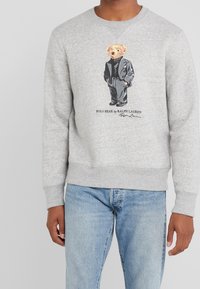 Grå sweatshirt med en Polo Bear-grafik i svart outfit, med ribbad mudd och fåll, tillsammans med ljusblå jeans.