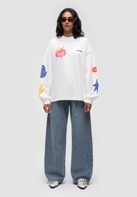 Camicia bianca a maniche lunghe con grafiche colorate; pantaloni in denim a taglio morbido; modello con occhiali da sole neri e scarpe dettagliate.