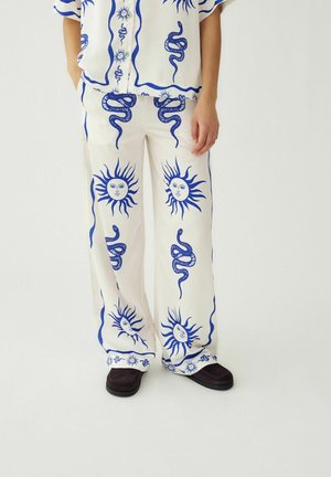 Pantalones anchos blancos con estampados de caras solares azules y serpientes enroscadas, acompañados de una camisa a juego y zapatos negros en una persona de pie.