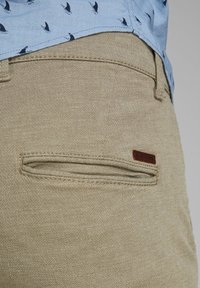 Khaki pants with textured fabric, featuring a horizontal zippered pocket and a small brown logo patch on the side seam.