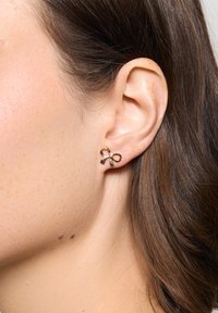 Złote metalowe kolczyki w kształcie kokardki, z małym centralnym kamieniem. Projekt zawiera pętle i akcenty w stylu wstążki.