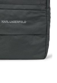 Geantă gri din nylon cu textură matlasată, având un buzunar frontal și logo-ul embosat „KARL LAGERFELD”. Formă rectangulară, cu închidere prin fermoar.