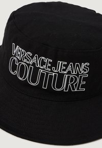 Crni šešir s ravnim rubom napravljen od teksturirane tkanine sa natpisom "VERSACE JEANS COUTURE" u bijeloj boji sprijeda, s ručno šivenim rubom.