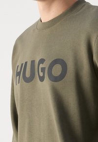 Olivově zelená mikina s kulatým výstřihem, vyrobená z měkké tkaniny, s velkým černým logem "HUGO" napříč hrudníkem. Texturovaný povrch.