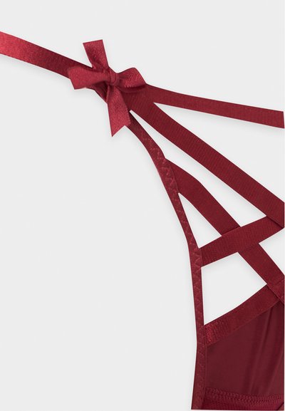 Gros plan de lingerie bordeaux à fines bretelles avec un nœud et des bandes élastiques fines qui se croisent sur un fond clair.