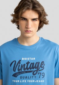 Šviesiai mėlynas medvilninis marškinėlis su apvaliu kaklu, kuriame ant priekio drąsiai atspausdintas juodas grafiškai tekstas "BIG STAR Vintage 79 Quality Your Life Your Jeans".