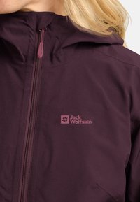 Giacca impermeabile viola con zip visibile e cappuccio. Presenta il logo Jack Wolfskin di colore rosa chiaro nella parte anteriore. Texture liscia.